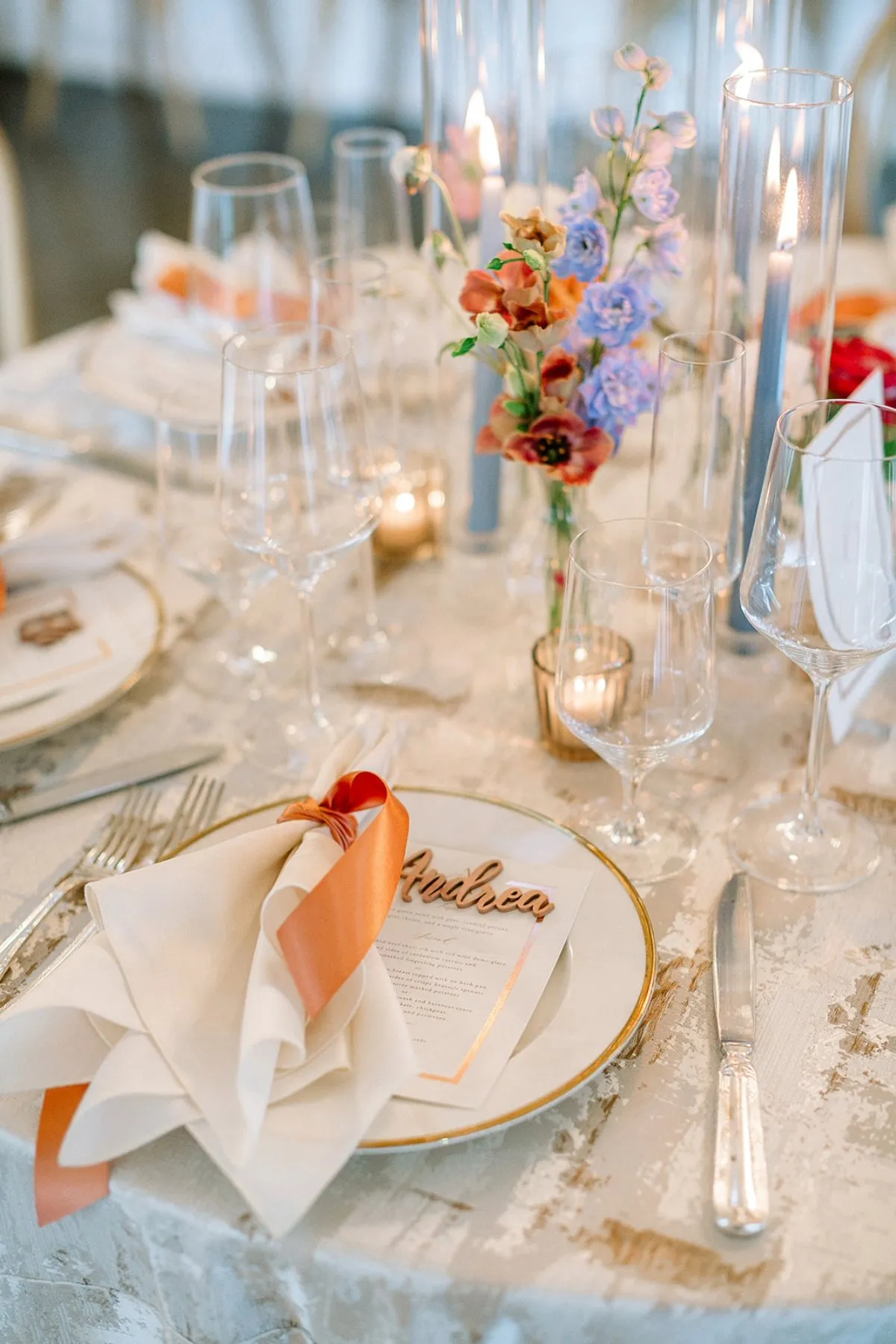 Pale custom-printed table linen with gold-rimmed china, orange ribbon napkins, clear glassware, colorful flowers, and candlelight. - Curated Linen Collection Pale custom-printed table linen with gold-rimmed china, orange ribbon napkins, clear glassware, colorful flowers, and candlelight.