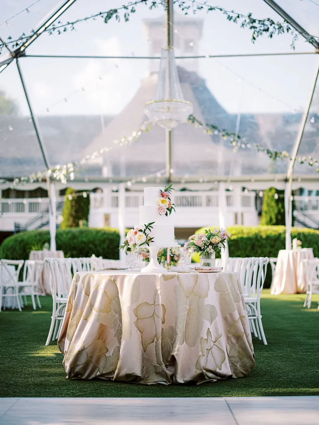 Wedding cake table styled with a cream floral linen beneath a clear reception tent. Wedding cake table styled with a cream floral linen beneath a clear reception tent.
