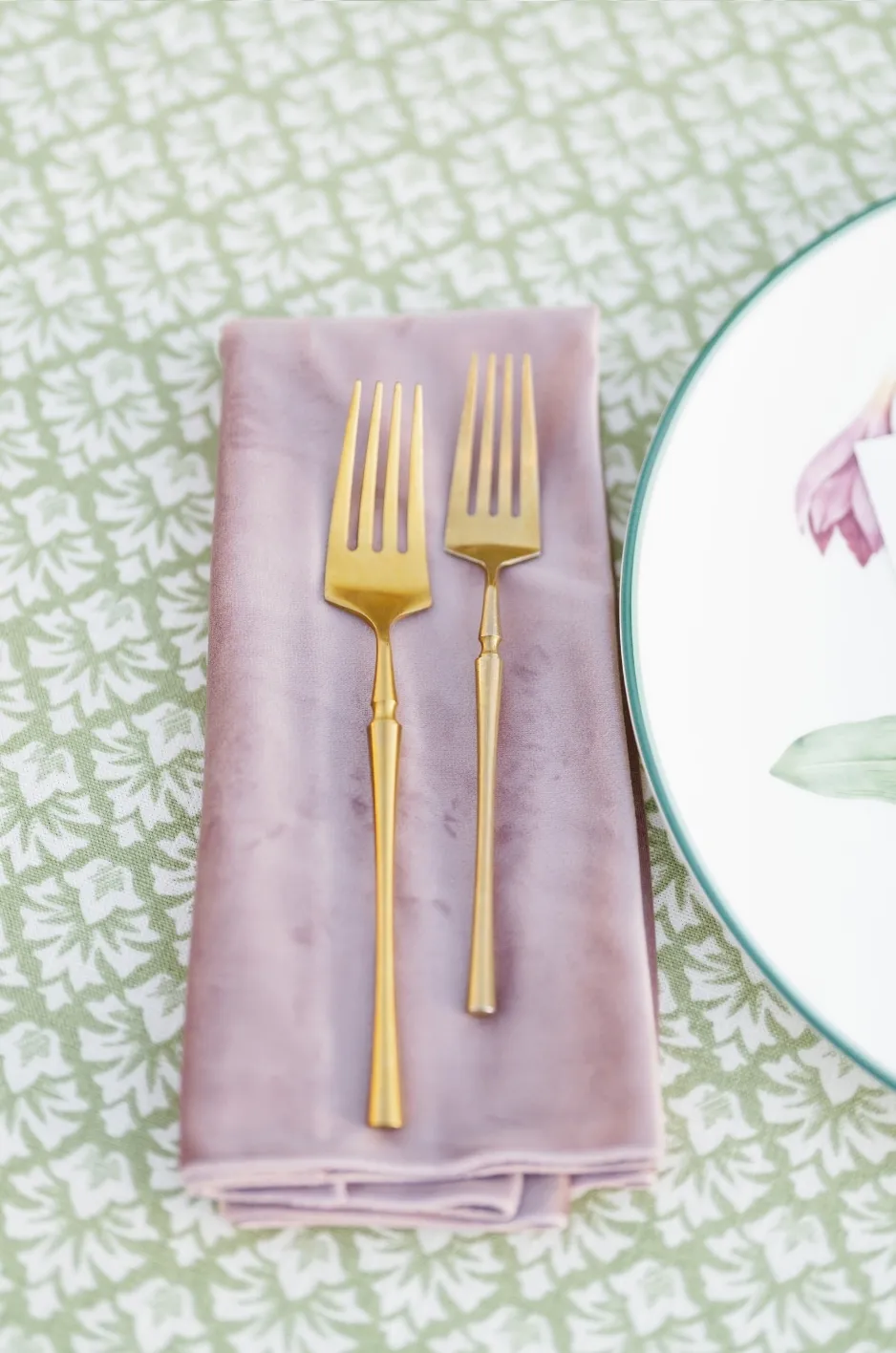 Folded blush napkin with gold flatware on a green patterned table linen - Curated Linen Collection Folded blush napkin with gold flatware on a green patterned table linen