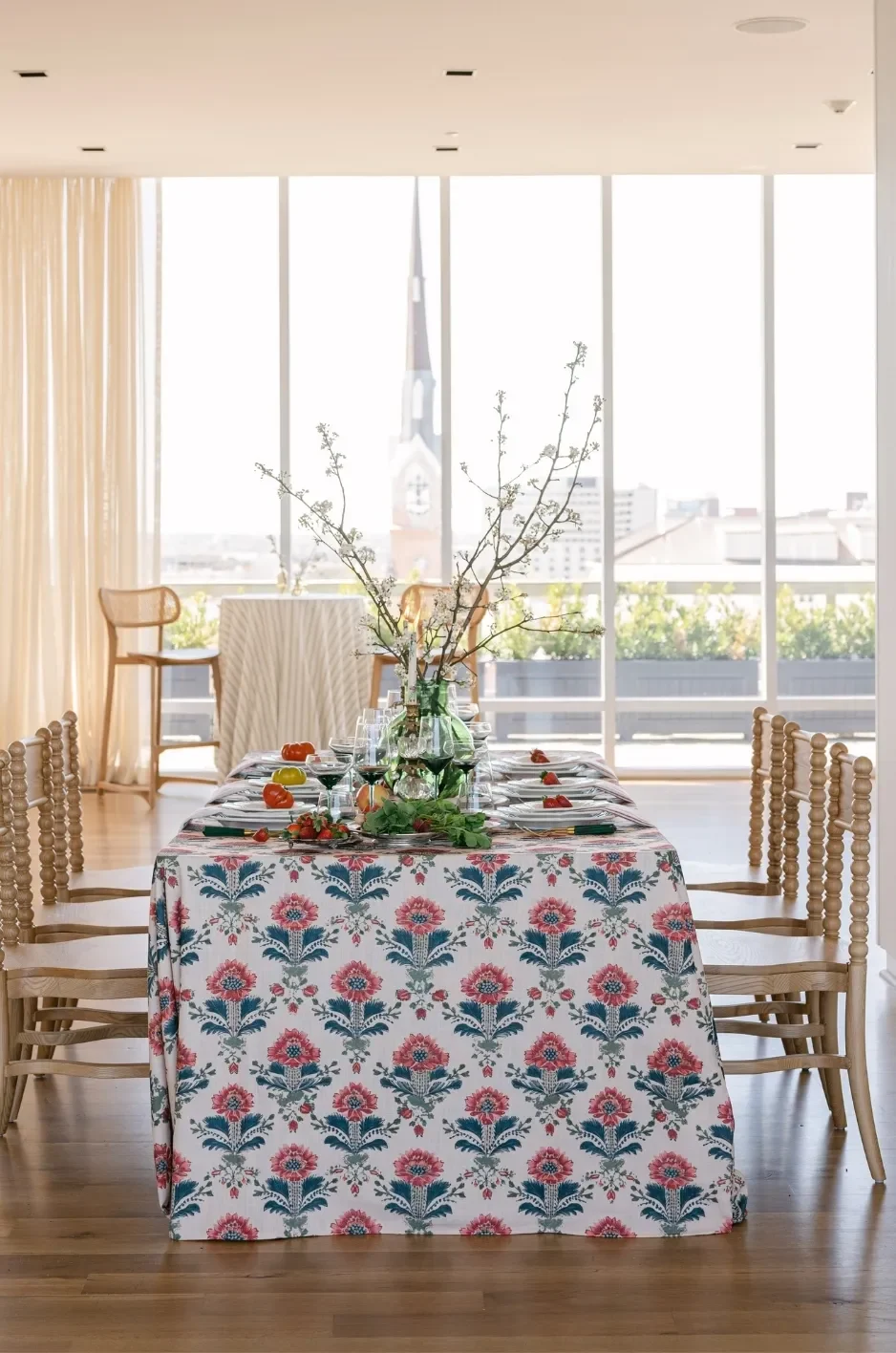Long reception table with a floral-patterned linen, branch centerpiece, and wooden chairs in a bright city-view venue. Long reception table with a floral-patterned linen, branch centerpiece, and wooden chairs in a bright city-view venue.