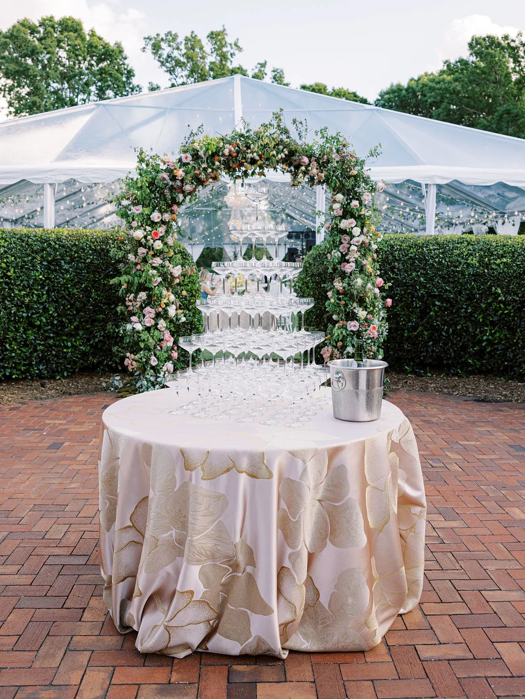 round table with pale floral linen and champagne tower beneath a floral arch at an outdoor tented event