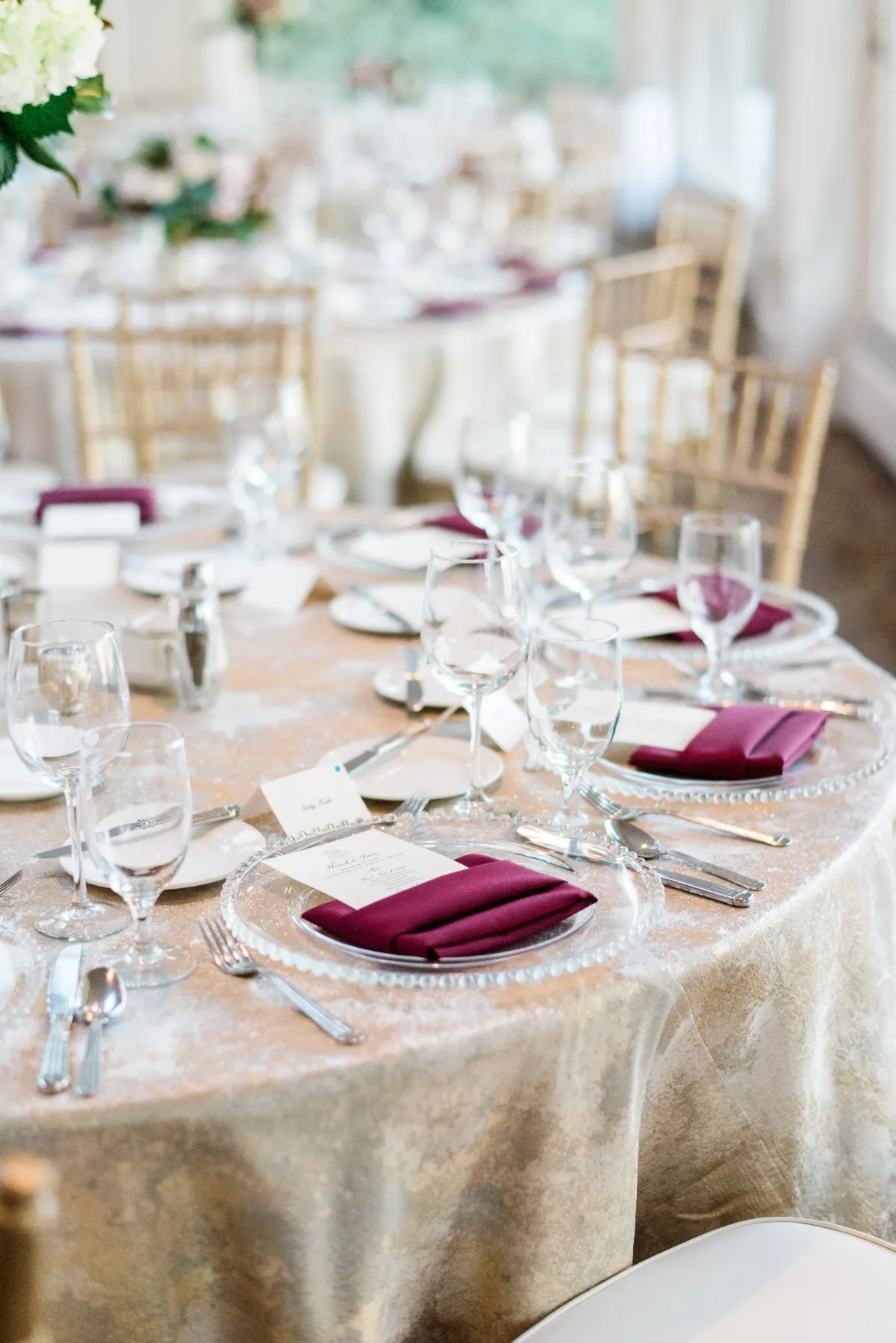 Plum napkins folded on clear chargers with white china, silver flatware, and glassware over a pale metallic linen. - Curated Linen Collection Plum napkins folded on clear chargers with white china, silver flatware, and glassware over a pale metallic linen.