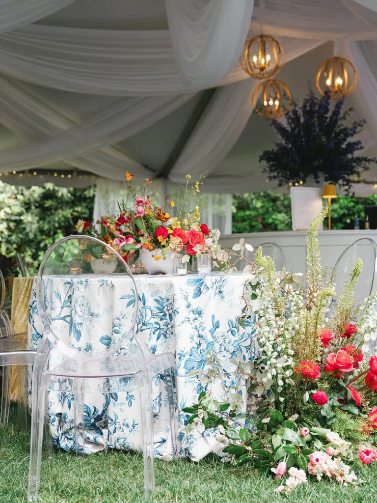Blue floral event linen with colorful flowers and clear chairs under a tented setting - Curated Linen Collection Blue floral event linen with colorful flowers and clear chairs under a tented setting