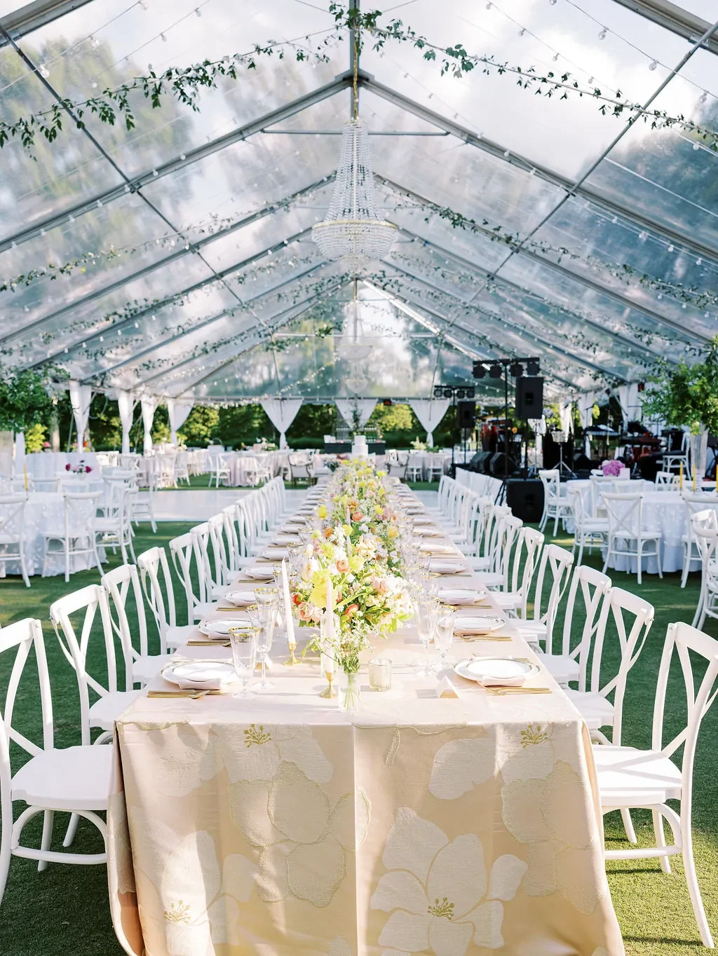 Long wedding table beneath a clear tent with a cream floral linen, white cross-back chairs, and garden-style centerpieces. Long wedding table beneath a clear tent with a cream floral linen, white cross-back chairs, and garden-style centerpieces.