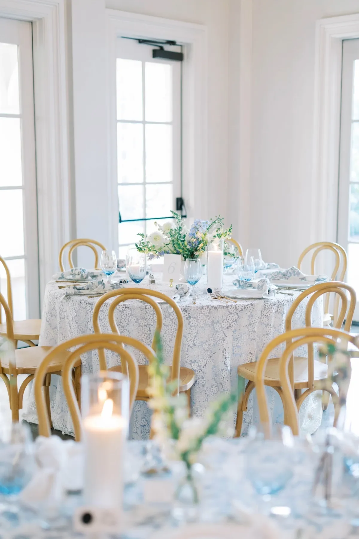 Round reception table with a pale blue floral linen, blue glassware, white florals, and natural bentwood chairs. Round reception table with a pale blue floral linen, blue glassware, white florals, and natural bentwood chairs.