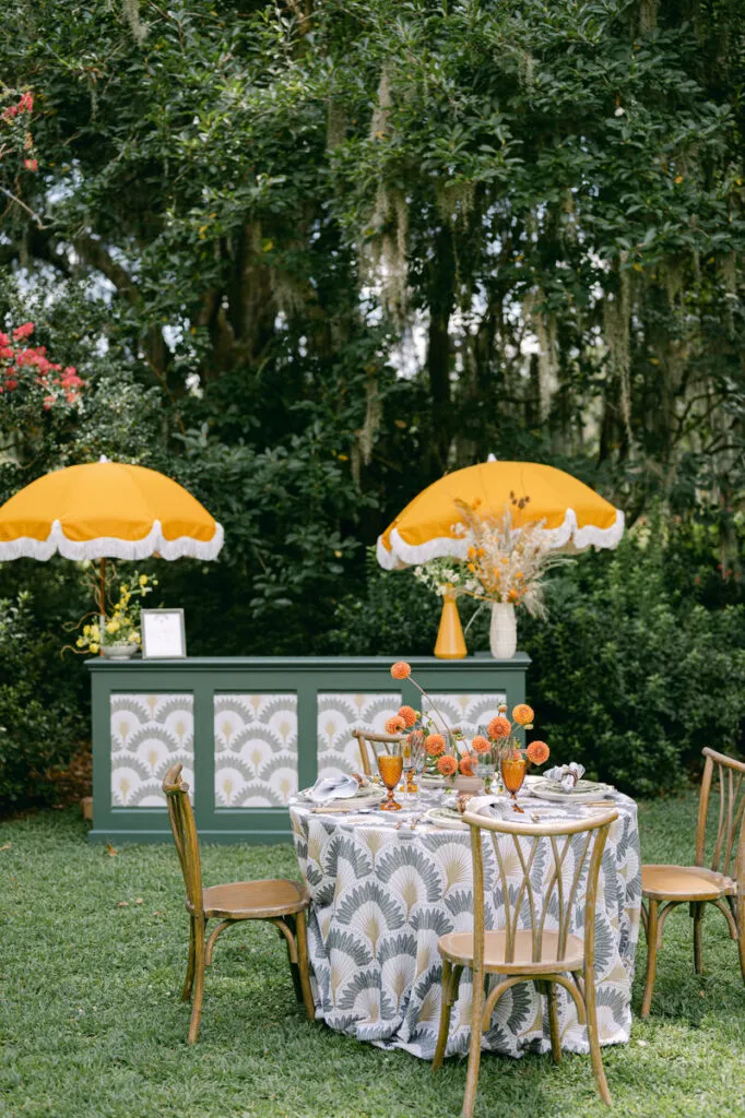 Outdoor event table with gray botanical linen, orange florals, green bar, and yellow umbrellas - Curated Linen Collection Outdoor event table with gray botanical linen, orange florals, green bar, and yellow umbrellas