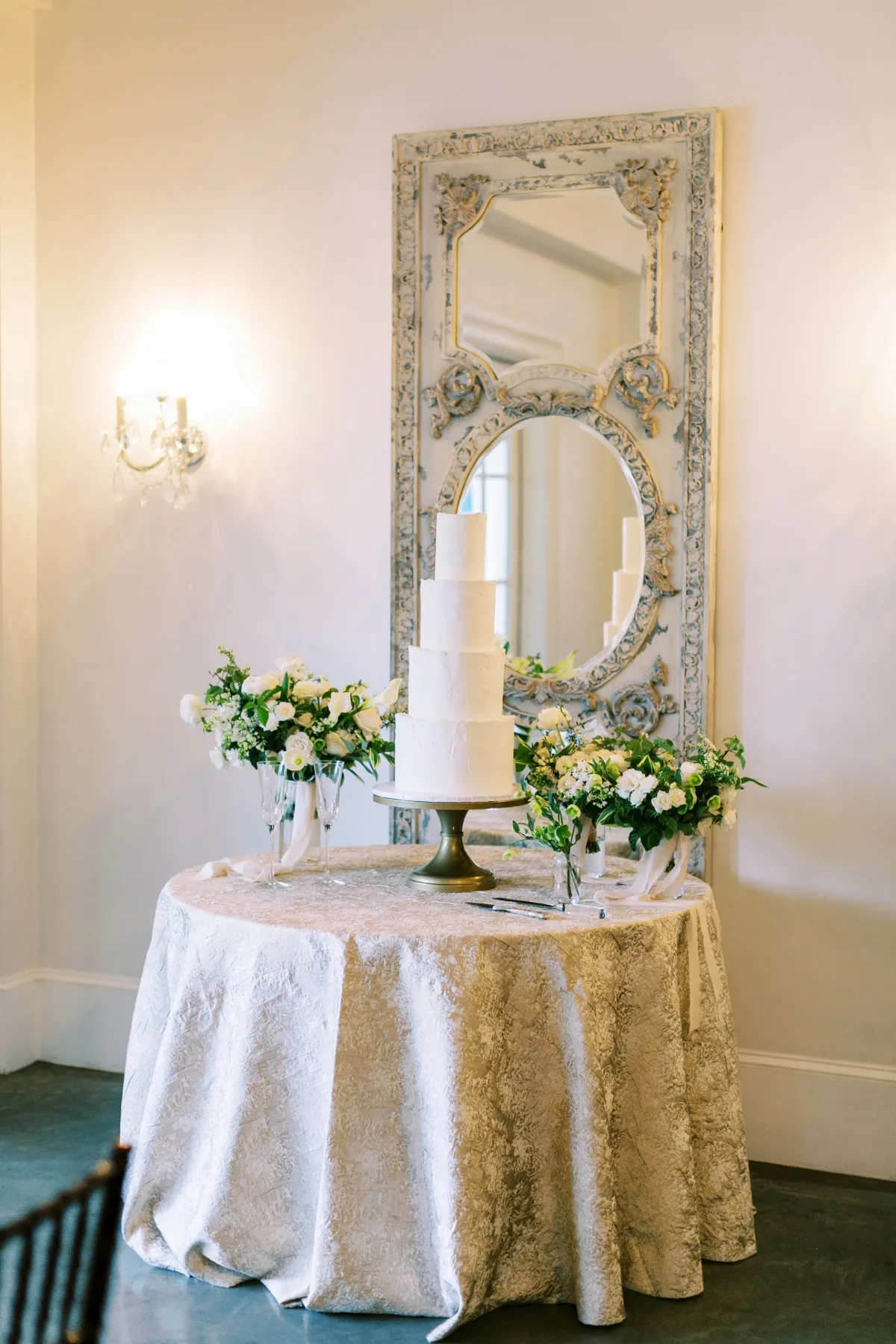- Curated Linen Collection Five-tier white wedding cake on a pedestal, flanked by white flower arrangements on a lace-covered round table in a room with an ornate, framed mirror in the background.