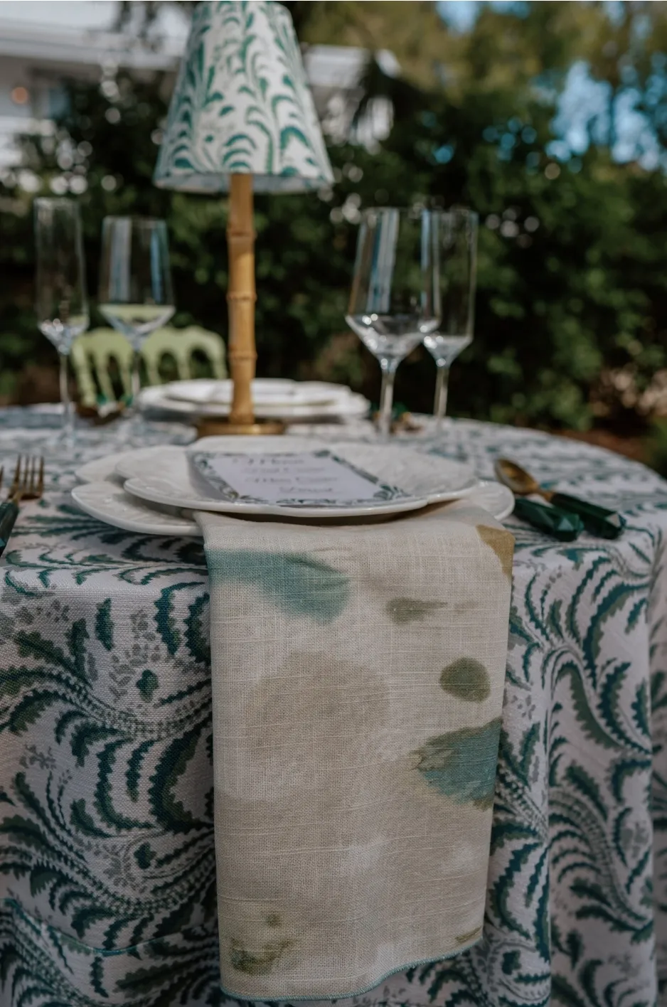Softly draped botanical napkin extending over a green-and-white patterned table with clear stemware and a shaded lamp. - Curated Linen Collection Softly draped botanical napkin extending over a green-and-white patterned table with clear stemware and a shaded lamp.