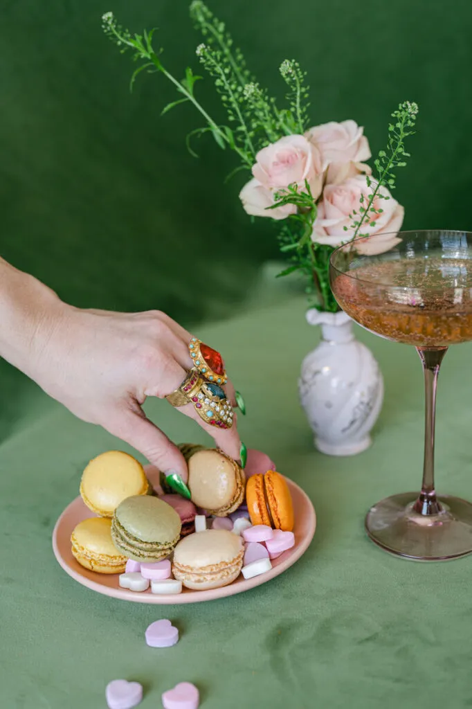 - Curated Linen Collection Hand with ornate rings reaches for pastel macarons on a pink plate, beside a vase of roses and a champagne coupe on a green table.