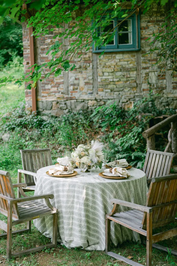 Outdoor stone cottage garden with a round table set for four, white patterned tablecloth, napkins, and a floral centerpiece.